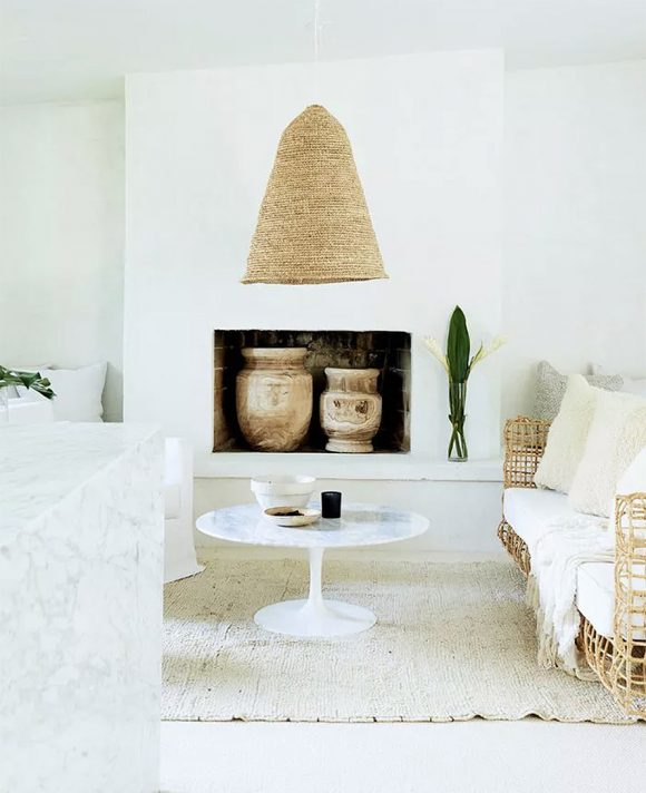 A beehive-shaped woven basket as a pendant light over a marble pedestal table on a sisal rug. Nearby is a plant-based woven sofa with 3 white embroidered pillows. In the fireplace behind, 2 large urns sit, while outside the fireplace is a plant frond in a clear vase.