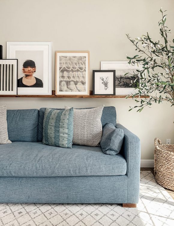 A ledge full of framed artwork floats above a blue sofa with grey and blue-green pillows, atop a latticework rug.