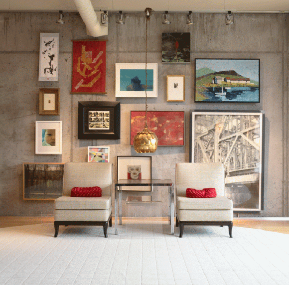 A large display of framed artwork in various sizes, colors, textures, and frame colors, hung with wire along a concrete wall, with a pair of beige slipper chairs and red back pillows between a table, under a gold pendant lamp.