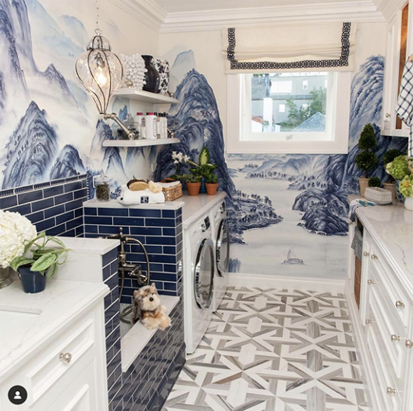 A white marble-topped sink, dark blue subway tiled dog-washing station, white washer and dryer beyond that, and matching white chest of drawers on the right in an award-winning mudroom at the San Francisco Decorator Showcase. On the walls is a mural in blue and white.