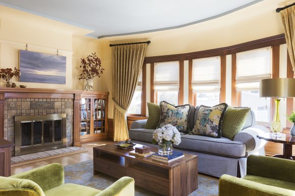 Living room with peachy walls, blue-grey ceiling, fireplace at the left with art above it and bookcase to the side, a periwinkle sofa with dark blue piping, 2 large pillows in lime green, and 2 large pillows in a blue, yellow, and white, floral with dark blue edging. Two lime green chairs are just out of view at the forefront, while in the rear, stacked and gathered gold spotted draperies flank a wall of 5 windows with wood edges and translucent blinds that fold up. A gold table lamp sits upon a side table, and a coffee table is dressed with books and flowers in the center.