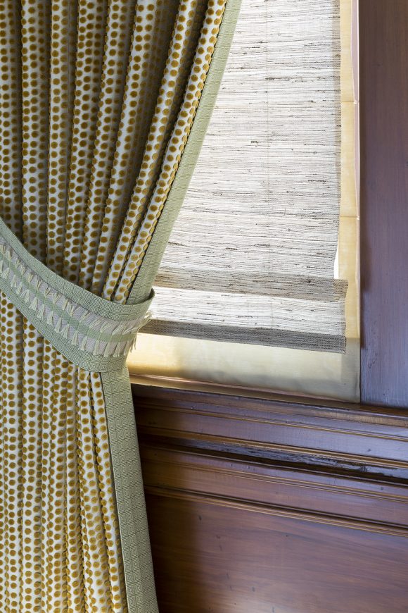 Close-up of a dark wood-edged window and stacked and gathered gold spotted drapes, showing edging in a moss green check, repeated as a tieback with repeating tassels in a lighter green. The natural grass blinds underneath have a textural quality, while a fabric liner at the back keeps light from spilling around, creating a soft light.