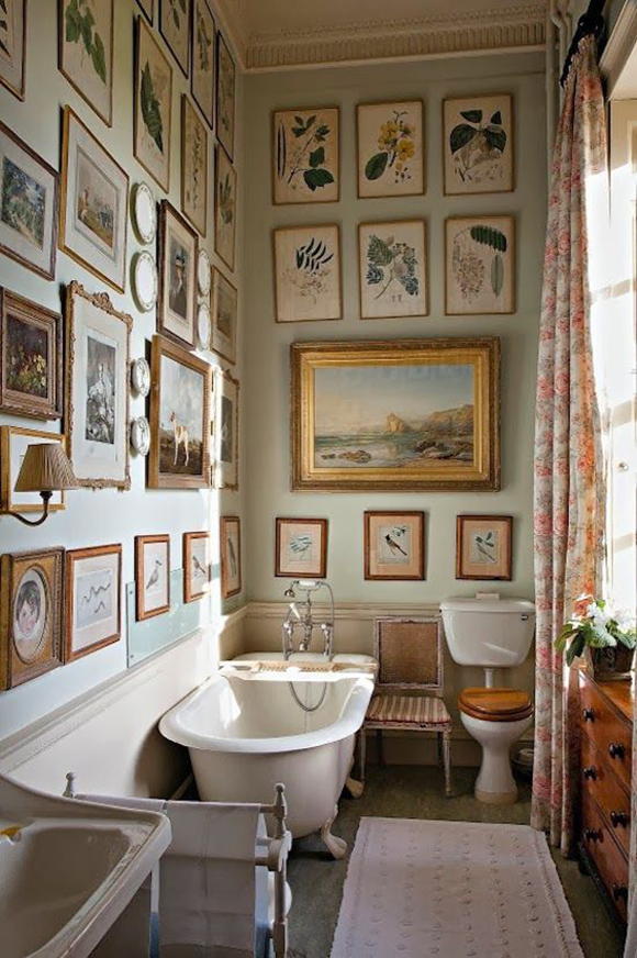 A white enameled claw-foot tub with an old-fashioned "telephone-style" faucet and spray nozzle sits underneath a series of neatly framed birds and landscapes on a soft green painted wall. Light streams in the open window dressed with floral curtains.