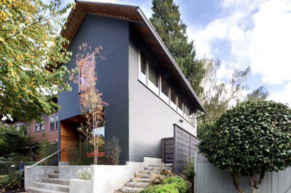 A grey wood-cladded ADU sits atop a set of concrete stairs, with clerestory windows along the side, and a warm wood entry space. Trees and green plantings make the building look like it belongs to the home it's attached to.