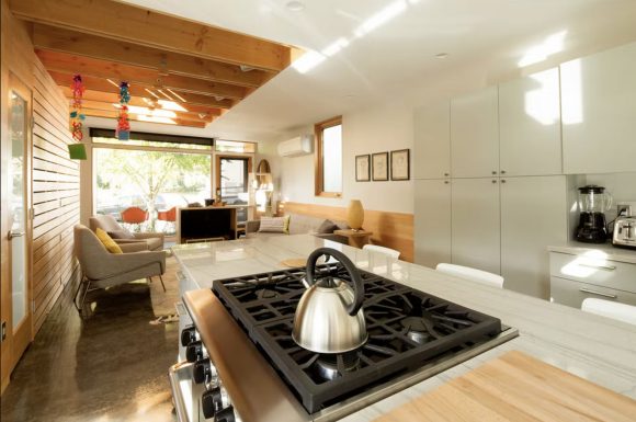 A long, narrow kitchen space is made wider with the use of warm wood paneling and light-colored cabinets. Light streams in from a large window at the end of the space that opens to a deck with outdoor seating. A set of clerestory windows brings in additional light that bounces around the cozy kitchen. A tea kettle sits on the cooktop surface in the foreground.