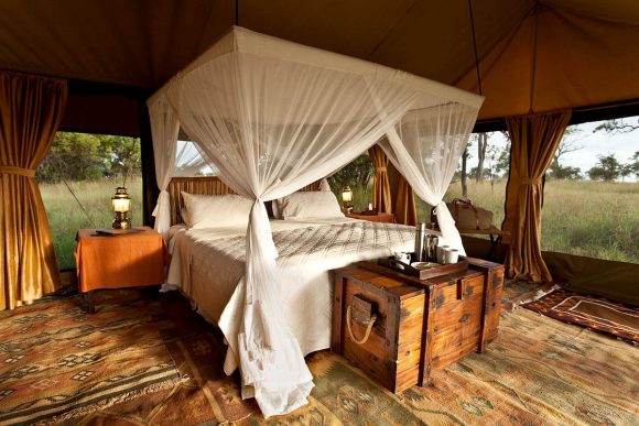 A canopy bed with mosquito netting tide to the posts has matching white blankets and sheets. At the foot of the bed is a wooden shipping box with rope handles, a coffee tray and two mugs upon it. The entire space is covered in tan and orange-y patterned rugs, while gold curtains are tied to the posts of the surrounding tent overhead. The view outside is green and hazy. A hurricane lamp sits on a bedside table covered in orange fabric, while a canvas duffel bag sits on another table on the far side of the bed.