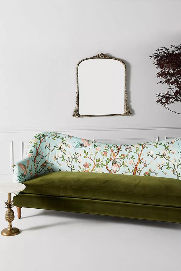 A sofa with a moss green velvet seat and winding multicolor floral fabric with a light blue background on the top half of the sofa sit on a plain white background, white floor against a white wall with a white chair rail. A simple metal frame mirror and a brass pineapple pedestal table with white marble top are nearby.
