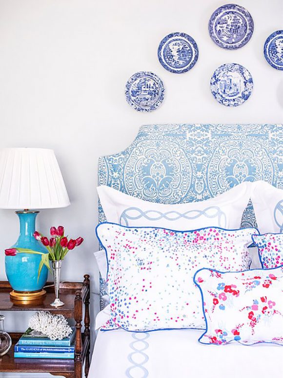 An upholstered bed with blue china- pattern-like headboard, covered in multiple white pillows with different types of florals and patterns, unified by the color blue. 5 blue and white plates are hung on the wall above the headboard. A stack of 3 shades of blue books sits on the bedside table, while a blue ceramic lamp and white lampshade sits on top. Red tulips in a silver vase finish the space.