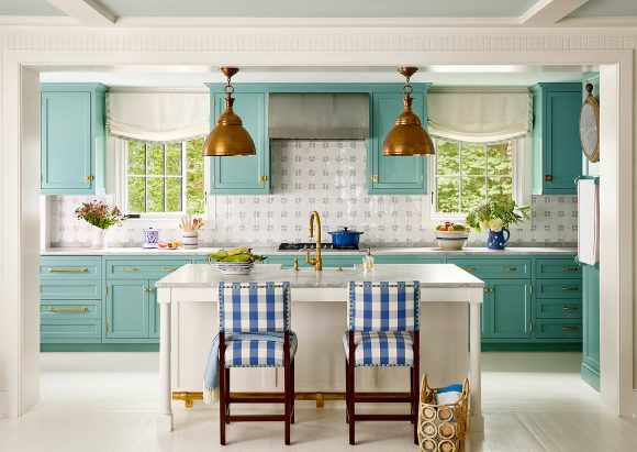 Head-on peek into a kitchen done in sea-green upper and lower cabinetry that has 4 units repeating a few feet apart and along the full white countertop length, brass cabinet pulls, and copper half-dish downlight pendant lamps. Blue and white checked gingham chairs are out front of a white kitchen island, while the square pattern is repeated in a grey and white backsplash.
