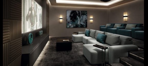 A tiered home theater room showing a bottom row of grey couches with turquoise pillows and side tables, and a higher row of turquoise couches with grey pillows. 3 sconces light up the back and side walls, white a coffered ceiling conceals white uplights. The projection screen shows on the left, facing the sofas, with sound baffling applied to the walls in geometric shapes.