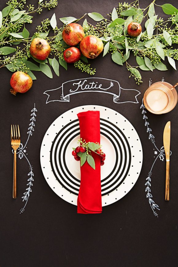 A table painted with black chalkboard paint has a white plate with dots and concentric circles set in the center, surrounded by drawn-on chalk leaves and a hand-drawn name ribbon. Atop the plate is a red fabric napkin rolled into a tube and fastened with sprig of red berries and leaves. Either side are a gold knife and fork, with a gold drinking glass above. At the top of the image, in the center of the table, is a layer of green leaves and stems, with red pomegranates scattered throughout.