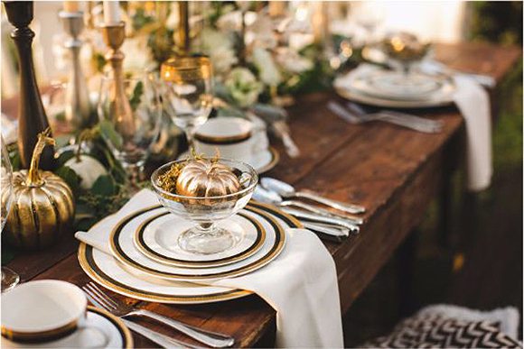 A traditional wooden table set with stacked plates in gold and black edging, with a glass pedestal bowl on top containing a gold painted mini pumpkin. 

Tall candleholders in gold with greenery around them and more place settings are out-of-focus in the background on the rest of the table.