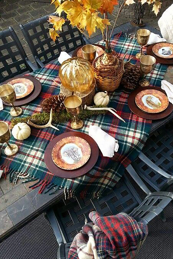 An outdoor dining table is set with a tartan throw blanket in green, red, and white plaid laid at an angle so that the fringe extends down either side on the diagonal. 

Plates are stacked with fabric napkins, set between gold metal stemware, while white mini pumpkins, a glass pumpkin in orange, natural pinecones with moss, and multiple stag horns adorn the center of the table.