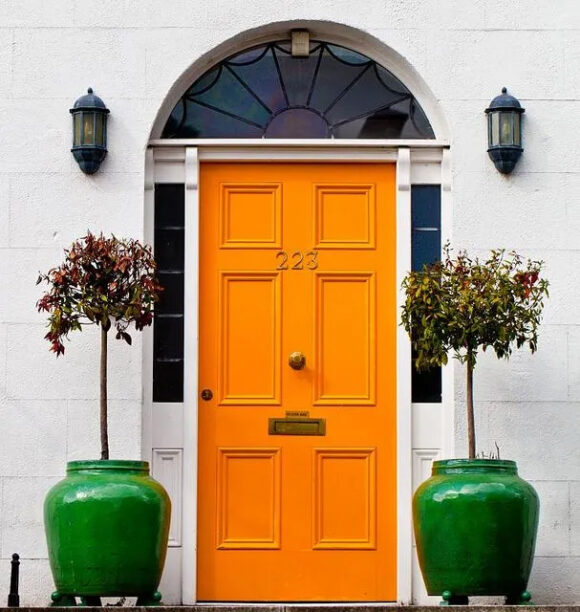 Bright orange front door between two glass highlights either side inset into a white brick wall, plus two small trees in oversize bright green pots flank the door. Two wall sconces are above the trees, either side of an arched window at the top. 