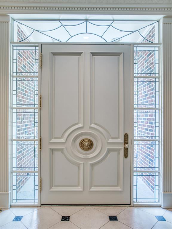 A large champagne-colored squareish front door with a circular handle in the center, and another more functional handle in the traditional spot on the right. A row of black-leaded side window lights balance the door, with a black-leaded spiderweb appearing to hang from the rectangular window above the door.