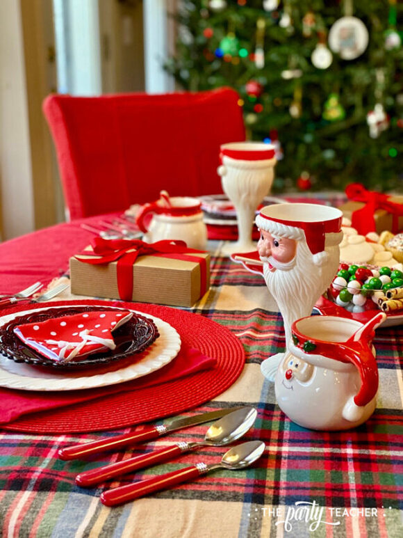 A red, green, black, and white plaid table runner underneath a red and white place setting with 2 Santa-shaped drinks containers, one tall and one short and round. A bowl of holiday-colored candies and a wrapped gift adorn the table, along with red handled silverware. A red dining chair and decorated Christmas tree are just out of focus behind.