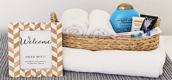 A framed sign with wifi login details, a woven basket containing shampoo, soap, toothbrush and toothpaste, with 2 small rolled white facial towels. The basket is sitting atop a larger white folded body towel.