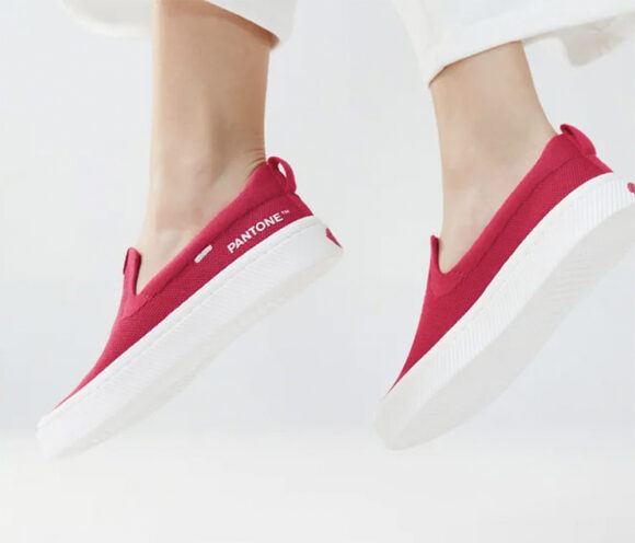 A pair of feet in slip-on shoes in the Viva Magenta color with thick white soles are jumping into the air against a white background.