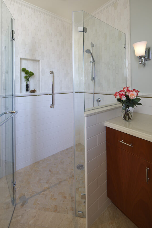 A corner curbless shower in white, and off-white wall tiles with tan and beige floor tiles at an angle. A clear glass door at the front. A clear glass half-wall at the top and tiling at the bottom separates the shower from the countertop and sink area.