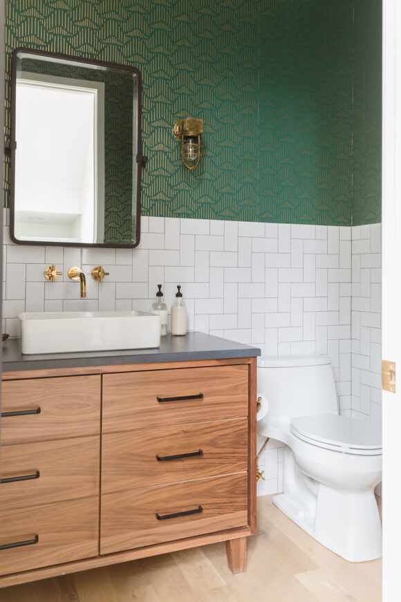 A bathroom's white 90-degree angled tiled wall sits just below a dark green textured wallpaper. Mirror, basin sink, brass wall taps, and a medium-light wood vanity with 6 drawers and horizontal black drawer pulls is beside a white toilet on blonde wood flooring.