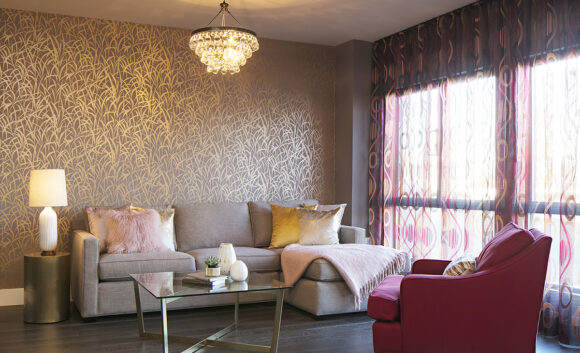 Gold textured wallpaper on the wall behind a purplish sofa with gold accent pillows and a glass coffee table, below a tiered ringed glass pendant light acting as a mini chandelier. The gauzy orange and hot pink draperies allow light to stream in from the two windows. A hot pink almost red armchair faces the purply-grey sofa. A side table holds a table lamp. The dark wood flooring grounds the space.