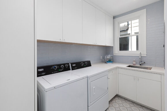 All-white washer and dryer appliances sit underneath white upper cabinets, and next to white lower cabinets, plus the under-mount sink and gooseneck faucet in silver.
