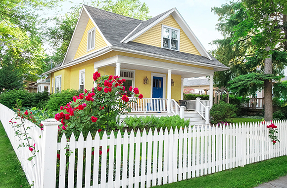 A yellow house with a gabled roof and white detailing around windows, doors, and porch railings that repeats on the which picket fence surrounding the green yard with a rosebush at the corner.