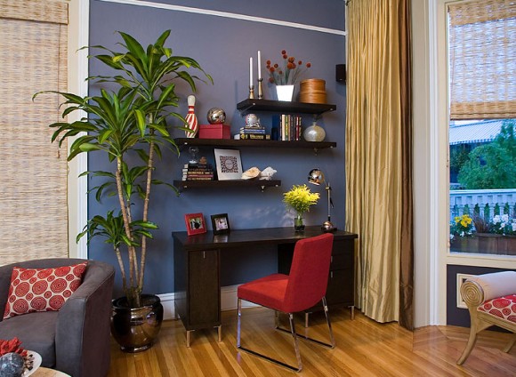 A home office in a corner with a saturated blue wall the shows off open shelving decorated with stacks of books, photos, candlesticks, vases of flowers, and other collectible items above a dark wooden desk with silver metal legs, a silver desk lamp, and a red chair with silver metal legs. A long gold curtain is gathered to the right which can be pulled across the windows on the right. A tall green spikey plant stands in a metallic circular pot to the left of the desk, and a brown tub chair with a lumbar pillow covered in red circles is just visible on the far left. A window seat upholstered in the matching red circle fabric is just visible in front of the right window. Beyond the brown chair on the left is a textural window covering that matches the window covering to the right.