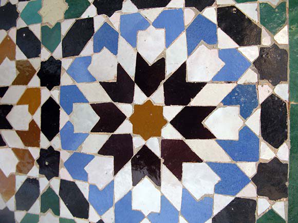 Closeup of geometric tiled shapes in concentric rings, starting in the center with a brick color 8-sided shape, then black arrows, then white interstitials, then blue arrow shapes, then black and green tiles.