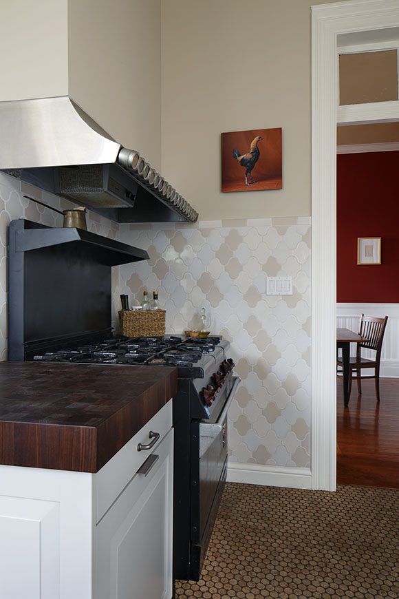 View of a farmhouse kitchen metal hood, range and cooktop, also showing tan and white tiling and wood butcher-block countertop end.