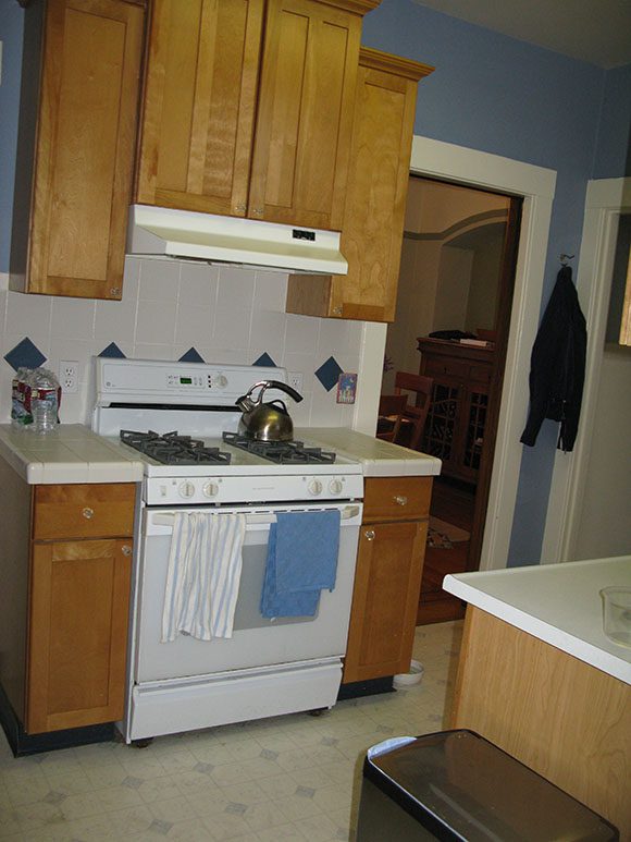 View of a white cooktop range and over, with a white standard hood over the top, and white backsplash tiling with a decorative green diamond trim that runs BEHIND the stove, and those orange wood cabinets surrounding. A doorway is to the right, and a peek of another countertop in the bottom right of the photo.