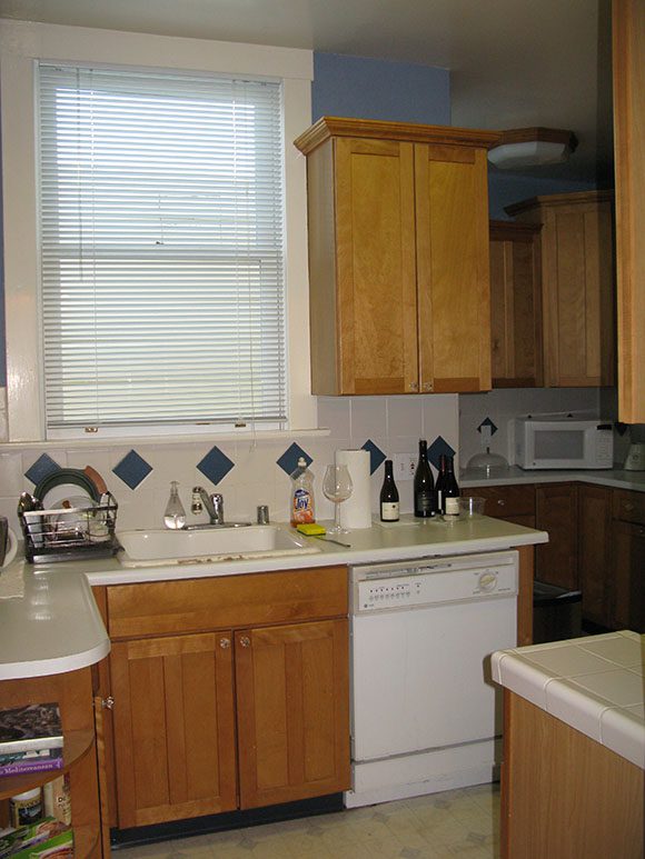 Photo of a dated kitchen, with a white plastic dishwasher front to the right of boring orange wood cabinets below an outdated sink with more cabinets wall hung above and to the right, and on the left, a large window, plus mismatched countertops.
