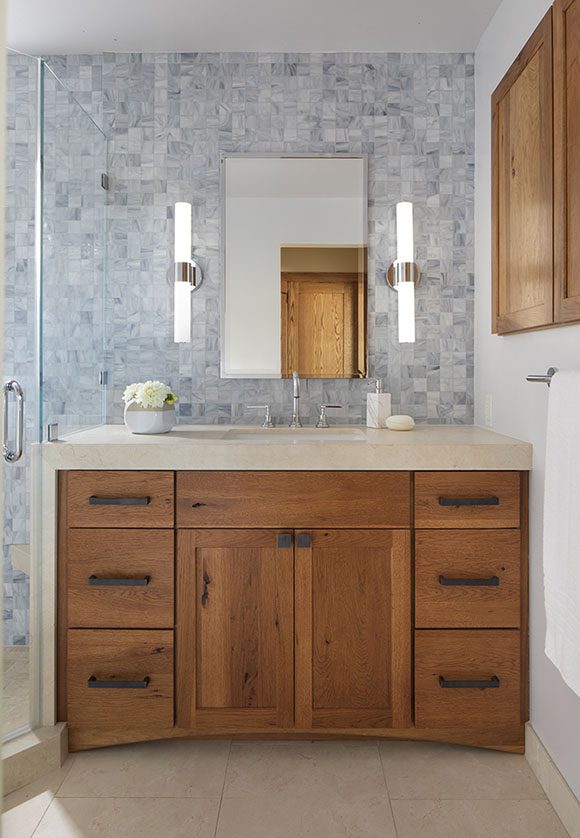 Head-on view of the sink and countertop in tan, warm wood cabinetry in an orangey brown with black metal hardware, mirror above with lights either side, walls in the same blue-grey mosaic tiling, bowed front along the bottom of the cabinetry, and tan marble floor tile, with towel rail to the right and a medicine cabinet in matching wood above that.