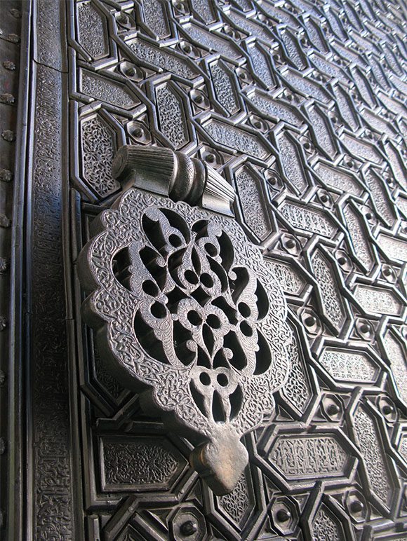 One large photo of a hand-beated metalworked door with a cutout door handle and repeating lattice-type pattern across the door.