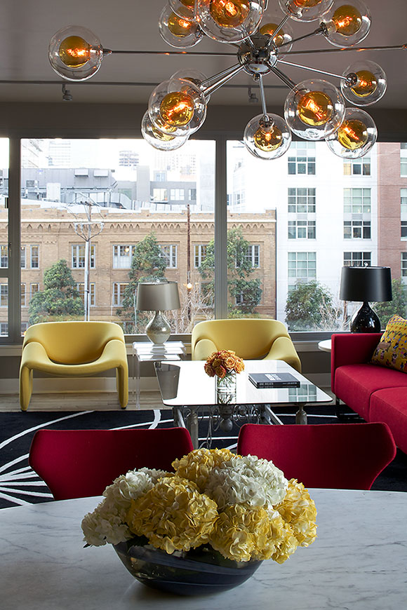 A bowl of yellow and white flowers adorn an Eero Saarinen marble pedestal dining table in front of two red B&B Italia dining chairs. Overhead is an amber bubble chandelier by Shades of Light. To the back are two modern yellow Artifort armchairs, a glass coffee table, and a red sofa.