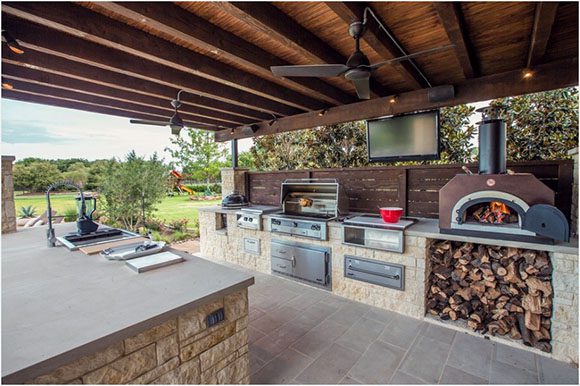 Backyard outdoor kitchen featuring overhead fans, rough stone island with smooth stone countertop, sink with spray nozzle. Across the stone flooring a few feet is a matching stone counter with BBQ grill, full oven and stovetop, warmer, and pizza oven, plus a ceiling mounted flatscreen TV, all with a view of the green grass and trees.