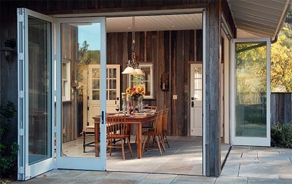 Exterior corner of a kitchen with folding glass walls stacked open, a single all-glass door remains in place. Wood cladding on the exterior complements similar finish on the interior, further blurring the distinction between indoors and outdoors. A reddish wood Stickley dining set with fresh flowers under a pendant light is the focal point.