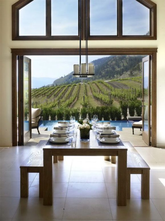A beautiful winery is framed by open folding double-width wooden and glass doors. In the foreground, a long oak table with benches either side is set for dinner guests, complete with fresh flower centerpiece. White outdoor chairs are peeking out from behind the edges of the open doors, while sunlight streams in.