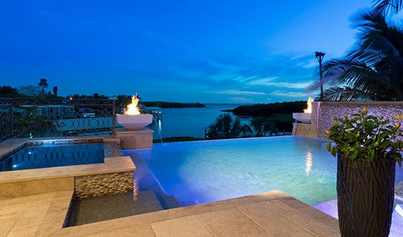 Pale flagstone surrounds steps into an infinity pool, with a hot tub accessible to the left. The infinity pool's edge is highlighted by two low columns topped with fire bowls. A planter and a palm tree show the right edges of our view, along a pebbled retaining wall. The darkening deep blue Florida sky with a just a brush of clouds shows above the line of the ocean for this exquisite backyard oasis originally designed by Bradenton Golf & Custom Pools, restored by Lucas Lagoons.