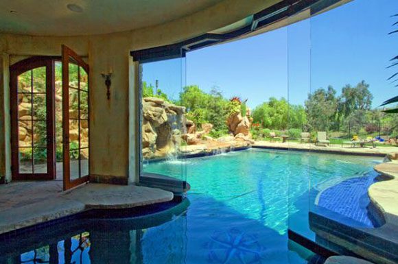 An unusual pool that begins indoors and extends outdoors, right through a wall! A pair of red French doors with one open inward show at the left, bounded by an aged, painted curved wall and ceiling that immediately becomes a huge folding glass wall, open at the center, then rejoining the solid wall. The tiled bottom of the pool invites you to try swimming between the indoors and out! The hot sun beckons, beating down on lush plantings and a large rock wall, with lounge chairs along the pool edge and pathway to the right.