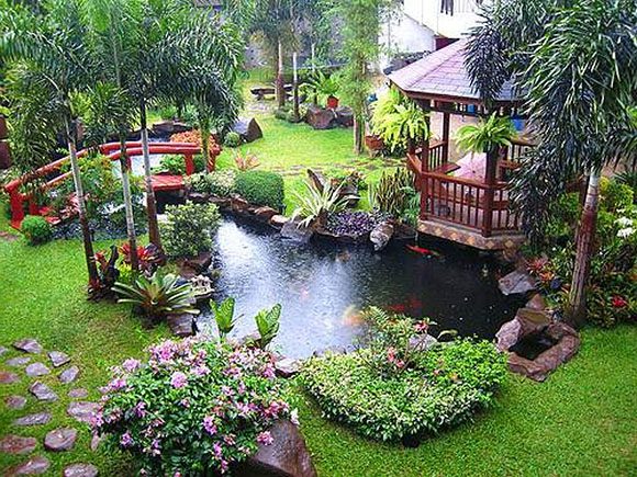 A traditional Japanese koi pond and backyard landscape design, surrounded by greenery, featuring a red bridge with a stone pathway leading to a gazebo with seating.