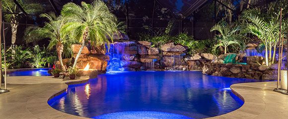 Night view of a Florida tropical backyard oasis of Zen calm, featuring a large curvy bright blue swimming pool, edged with light colored large flagstones, surrounded by multi-level rock walls, palm trees and lush plants. The Zen wading pool peeks out the left, just behind a fire pit and tree by the emerald blue rock waterfall, grotto, and hanging gardens. Uplit plants and trees throw interesting shadows.
