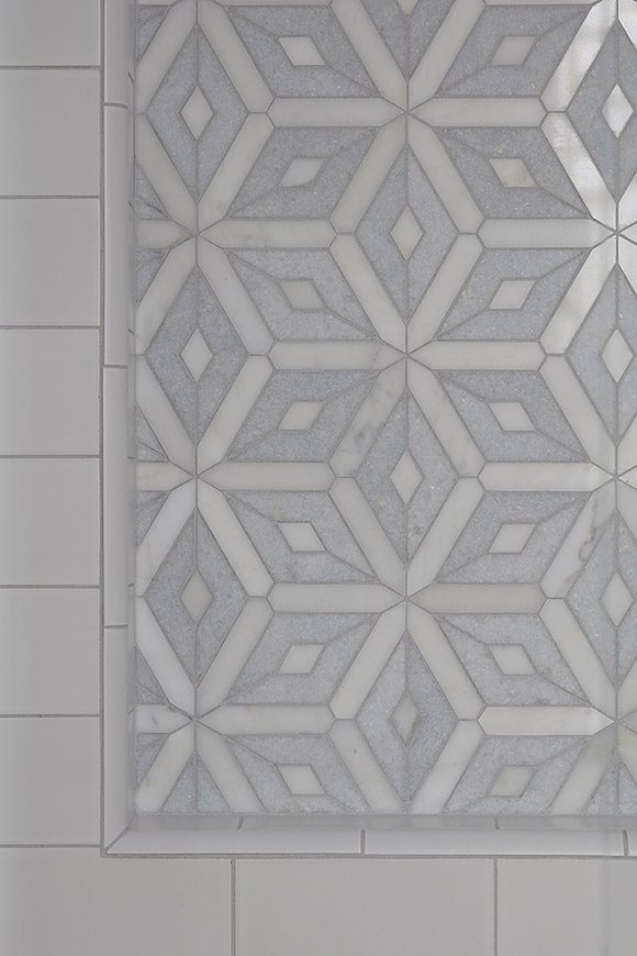 Closeup of shower wall inset niche, pattern created in pearly white Calacatta marble, shaped almost like snowflakes, alternating with speckled pale blue diamond-shaped Celeste marble, framed by off-white subway tiles.