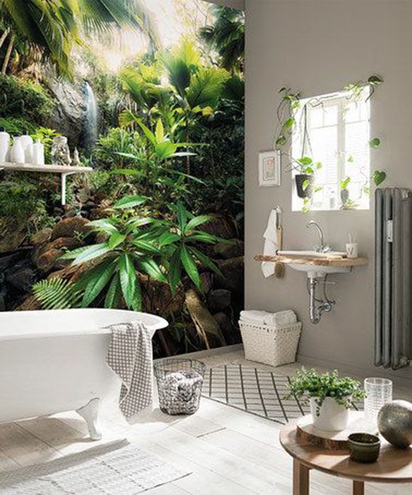 A Bohemian bathroom with a white claw-foot tub on the left, in front of a full wall mural of lush green plants and a waterfall, and to the right, a grey wall with wooden sink surround below a windowsill circled by living vines. On the floor a white woven basket of clean towels rests near a lattice-design rug, a wire basket for dirty towels, and to the far right a wooden table with a potted plant and some collectible glass and stone items.