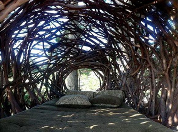 The view out, seated inside a human-scale nest, created by folded tree branches. A few pillows on a mattress, scattered beneath dappled light, mostly shaded by the branches, created by artist Jayson Fann.