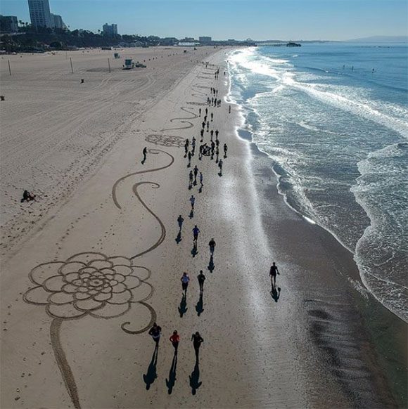 Multiple geometric shapes at enormous scale are created on the beach, using only brushes on sand and water, by Artist Andres Amador. In this view, the ocean is on the right and the sand is on the left, and we're looking all the way down the beach, while tiny people walk past.