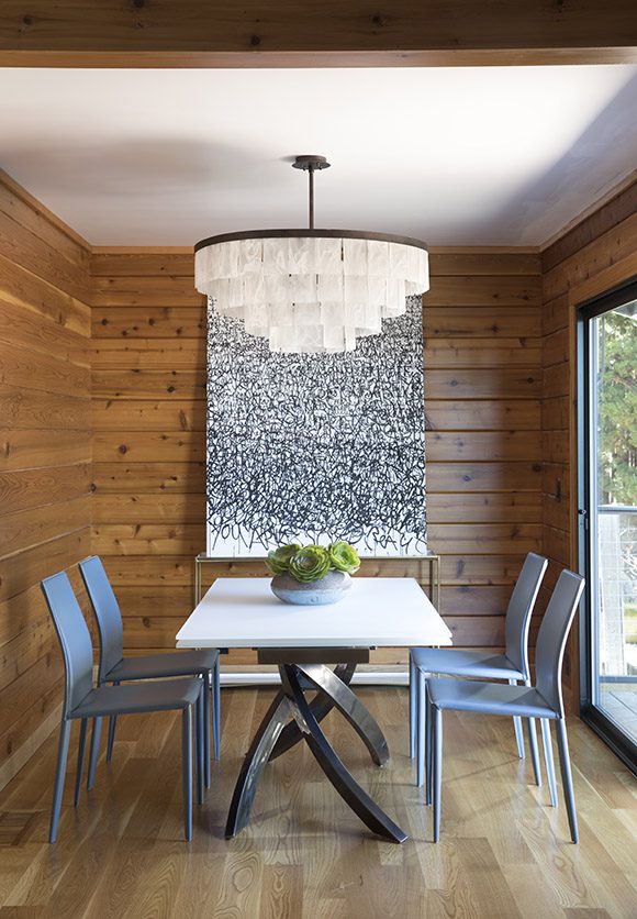 Close-up of a contemporary dining room showing white glass elements on the statement-piece round chandelier, an extendable white glass dining table, leather dining chairs in a light blue color, and a black and white patterned artwork hung on the back wall. A sliding glass door to the right of the image brings light into the space, while the knotty cedar wood walls feels cozy and natural.
