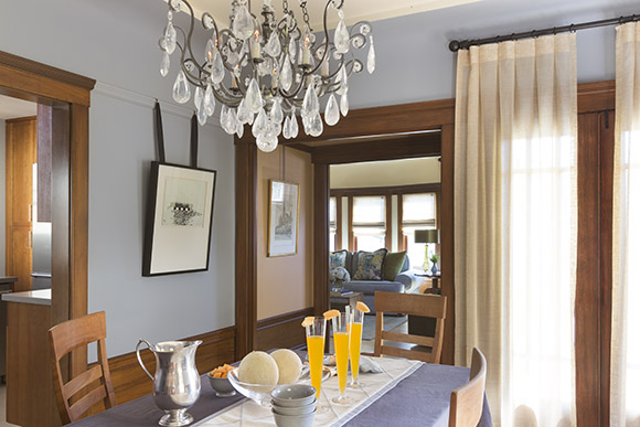 Traditional dining room featuring wood trim and wainscoting. Artwork is hung from a picture rail with black grosgrain ribbon. A Schonbeck rock crystal chandelier hangs above the dining table, covered with a blue table cloth and set for breakfast. Light is filtered through full-length sheer curtains with a box pleat on the right. Straight ahead is the doorway into the foyer, and to the left is the doorway to the kitchen, all finished in warm wood trim. Three glasses with orange cocktails sit in the center of the table, next to a bowl of melons, and a silver pitcher of water.