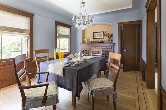 Dining room, showing a table with tablecloth under a Schonbeck rock crystal chandelier, custom seat pads on 4 wooden chairs, natural grass woven roman window shades, wood flooring and wood paneling with sky blue wall paint color and a melon-colored arched wall niche with a wooden storage unit.