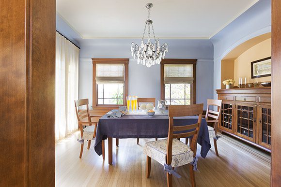 Looking into a traditional dining space in the style of Americana or historic arts and crafts, shown by the wood flooring, wood trim, wainscoting and picture rail. The recessed ceiling tray is painted a crème color, to differentiate it from the curved cove painted in melon color and the walls in a sky blue. A rock crystal chandelier hangs above the dining table, covered with a blue table cloth, and the wooden chairs have custom padded seat covers in a light-colored pattern. Roman shades in off-white finish the wood-trimmed windows, while a wooden storage unit is tucked into the arched alcove to the right of the space. Light is filtered through full-length sheer curtains on the left.